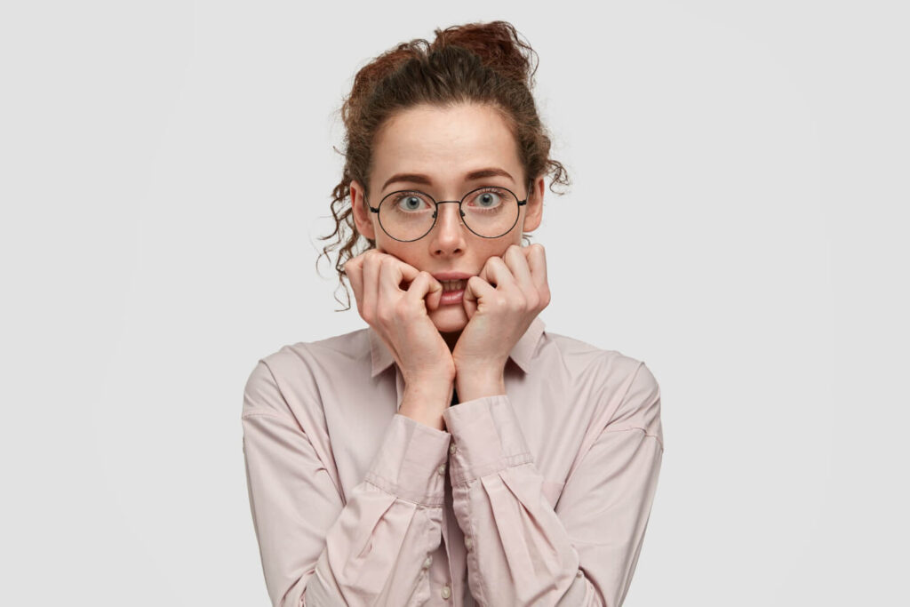 unhappy nervous young female with worried expression bites finger nails looks anxiously directly wears spectacles dressed fashionable clothes stands against white wall 1